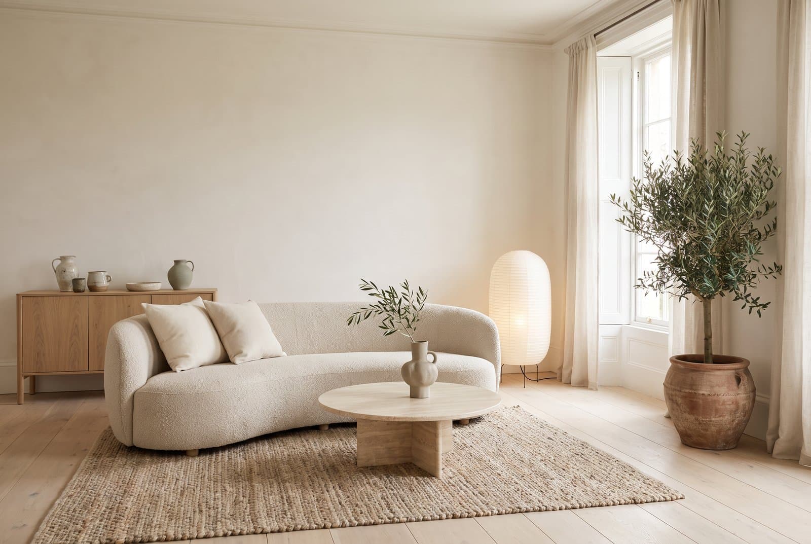 Concept visual of a calm organic modern living room with warm white walls, wide pale oak floorboards, a curved cream boucle sofa, a round travertine coffee table with a stoneware vase of olive branches, a chunky jute rug, a paper floor lamp, an oak sideboard with handmade jugs and an olive tree in a terracotta pot by a tall window with linen curtains
