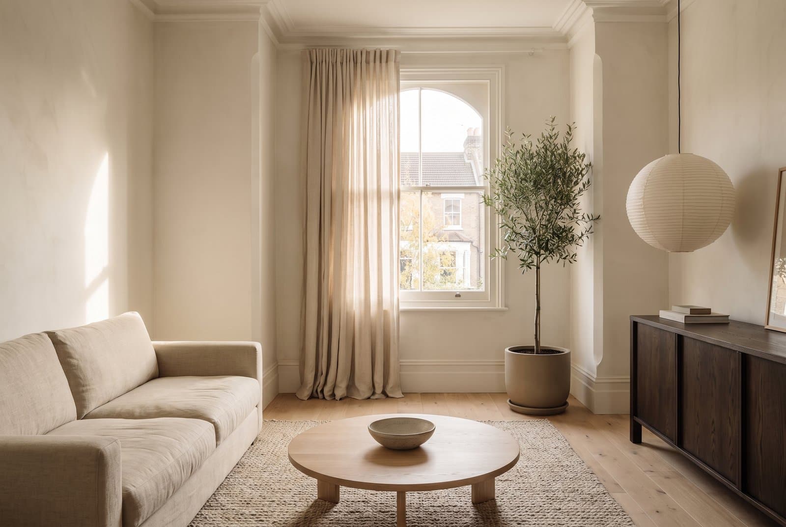 Concept visual of a warm Japandi living room in a Victorian terrace, with chalky limewashed walls, a low oatmeal linen sofa, a round pale oak coffee table with a stoneware bowl, a woven wool rug, a dark wood sideboard, a paper lantern and an olive tree by a tall window with linen curtains