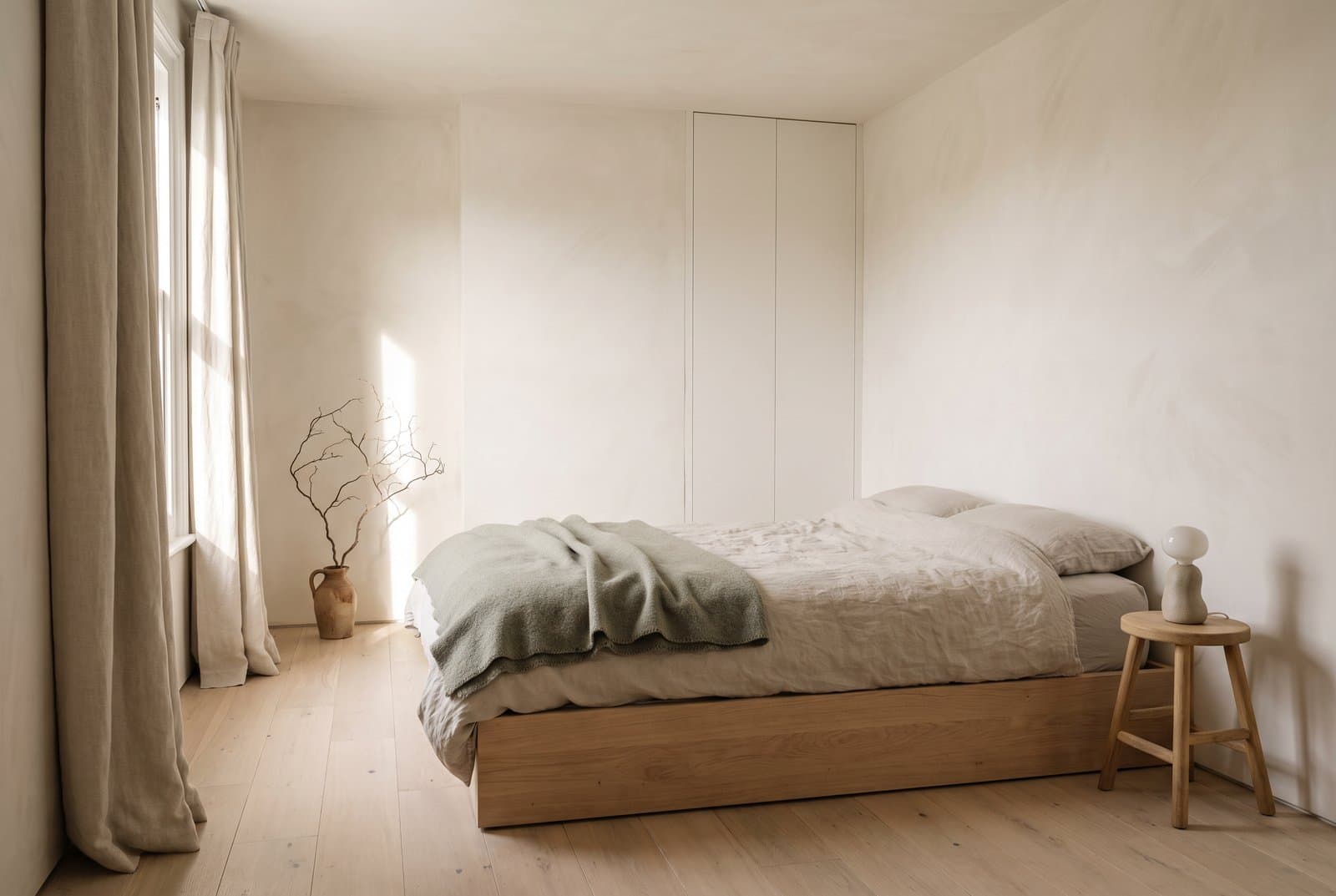 Concept visual of a warm minimalist bedroom with chalky limewashed walls, flush built-in cupboards, a low oak bed in creased oat linen with a sage wool throw, an oak stool with a stoneware lamp, linen curtains and a bare branch in a clay jug