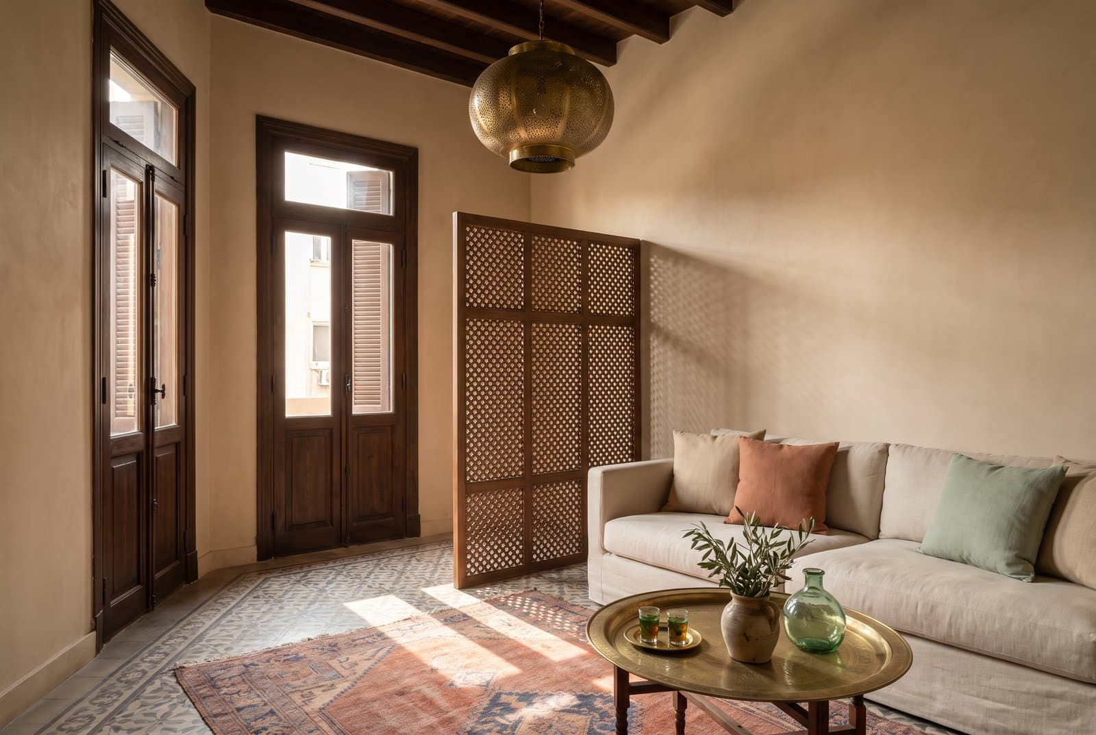Concept visual of a calm Cairo apartment sitting room with warm sand plaster walls, dark timber beams, tall shuttered double doors, a wooden lattice screen casting patterned shadows, a cream linen sofa with oat, terracotta and sage cushions, a pierced brass lantern, a faded rust rug on patterned floor tiles and a brass tray table with two glasses of tea, an olive branch and a green glass bottle