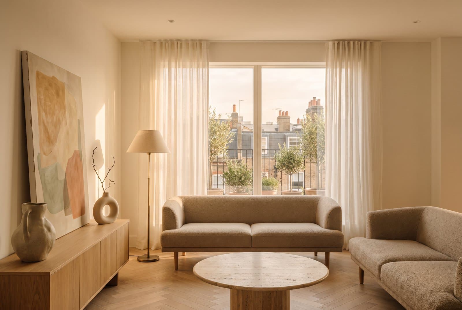 Concept visual of a warm urban chic living room in a city flat at golden hour, with a low stone-coloured sofa, a round travertine coffee table, a brass floor lamp, a pale oak herringbone floor, sheer linen curtains, a large abstract painting in earth tones and a balcony of potted olive trees overlooking the rooftops